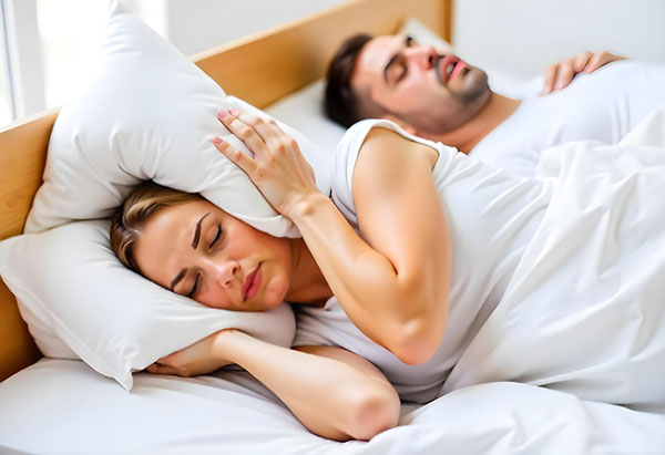 Woman covering her ears with a pillow while sleeping next to a snoring man in bed